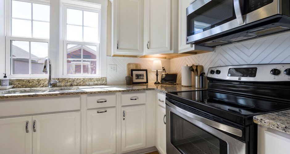 Bright white kitchen with granite counters, stainless steel stove and microwave, and large front windows.