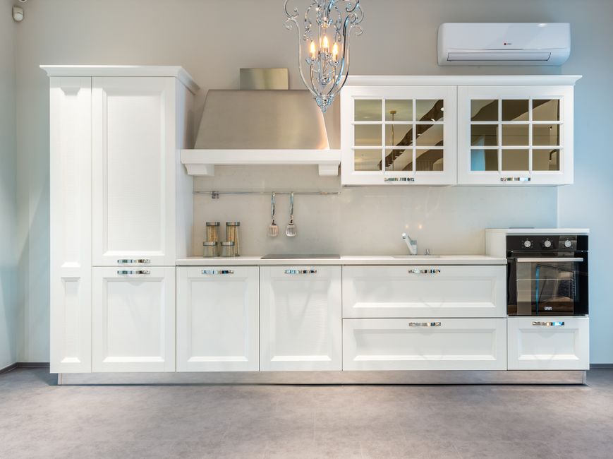 White modern kitchen with tall cabinet, stainless range hood, glass-front upper cabinets, built-in oven, and a chandelier above."related to kitchen design"