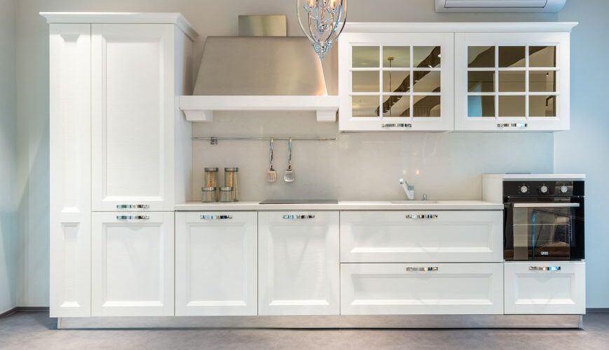 White modern kitchen with a tall cabinet, stainless range hood, glass-front upper cabinets, built-in oven, and a hanging chandelier.