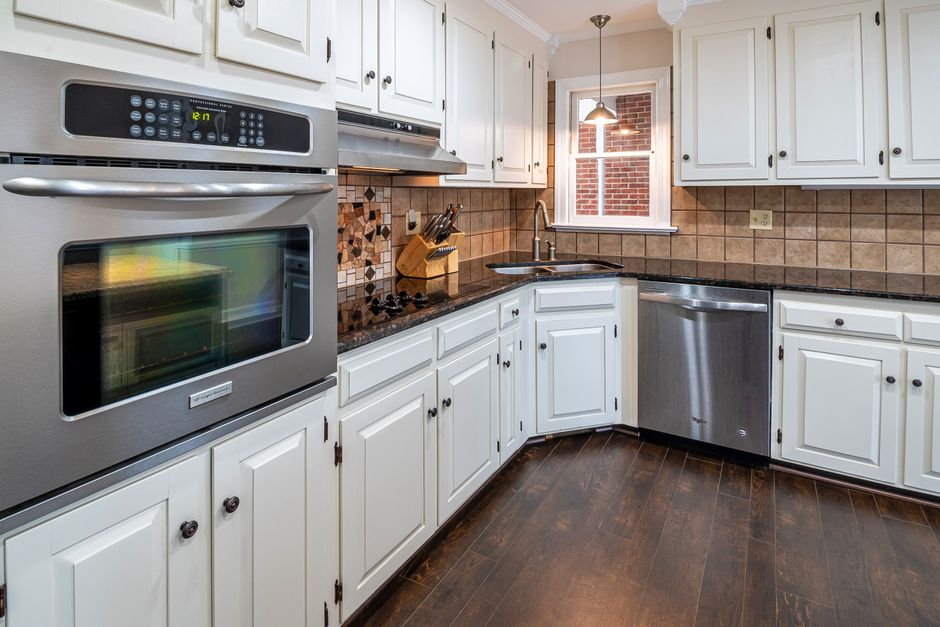 Modern L-shaped kitchen with white cabinets, stainless steel oven and dishwasher, and dark hardwood floors.