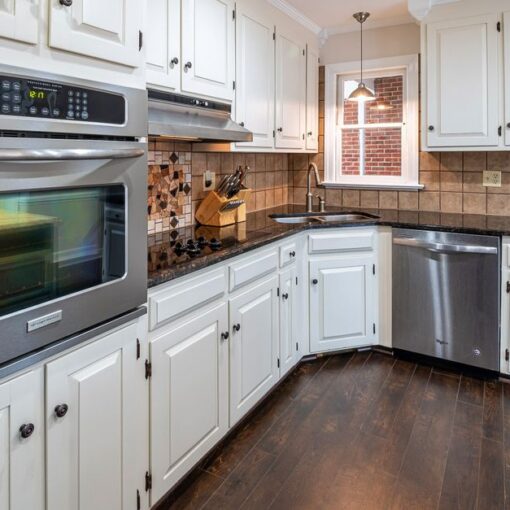 White kitchen with stainless steel double oven, dishwasher, and dark wood floors; tiled backsplash and window above sink.