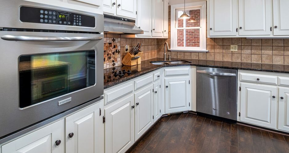 White kitchen with stainless steel double oven, dishwasher, and dark wood floors; tiled backsplash and window above sink.